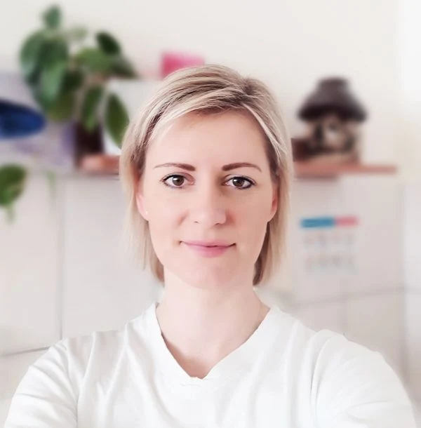 Žena v bílé košili se usmívá na pozadí s rostlinami.
