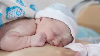 Spící novorozené dítě v bílé čepici na rukou dospělého.