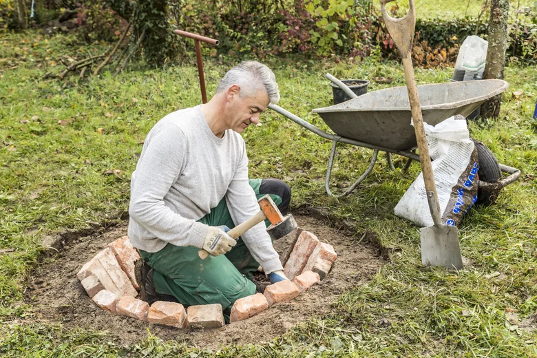 Muž staví kruhové ohniště z cihel v zahradě.