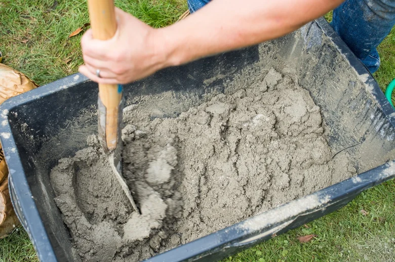 Člověk míchá beton lopatou v plastovém voze na trávě.